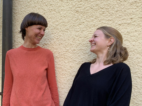 Die zwei Geschäftsführerinnen von Mokä, Ina und Fabienne, stehen vor einer beige strukturierten Wand und schauen sich fröhlich an.